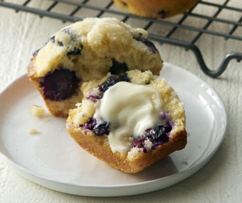 A blueberry muffin cut in half with butter on one half on a plate