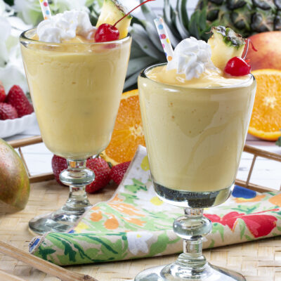 Two glasses filled with yellow blended beverage garnished with whip cream, pineapple, and a cherry