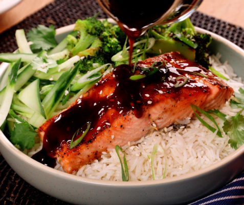 A bowl filled with rice topped with salmon, mixed green vegetables and a drizzle of teriyaki sauce