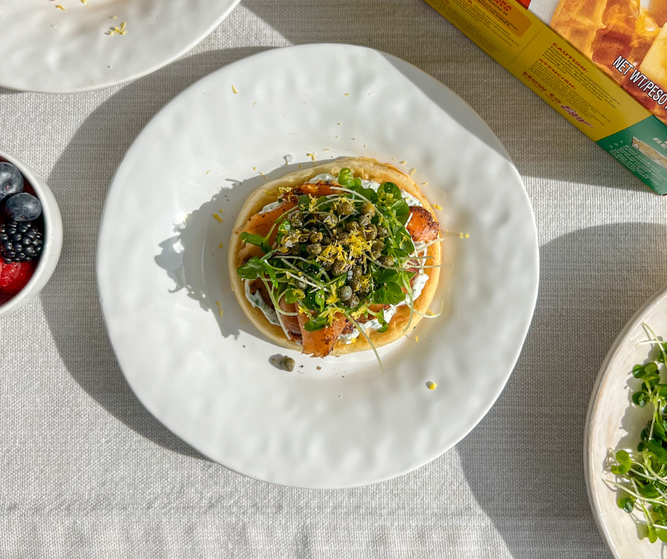 a waffle topped with smoked salmon, cream cheese, and various greens on a plate