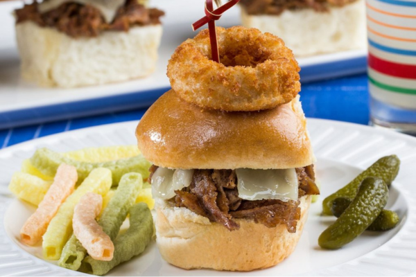 A cheesy steak slider with an onion ring on top on a white plate next to veggie straws