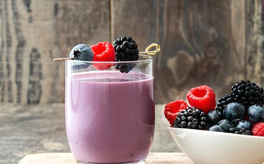 Purple smoothie next to a bowl with raspberries, cherries, and blackberries in front of a wooden background