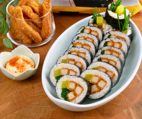 A long plate with 2 lines of chicken kimbap next to a sauce cup with dipping sauce and a glass with chicken fingers