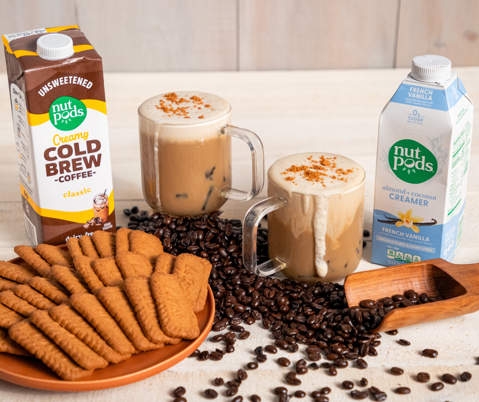 Two mugs filled with coffee surrounded by coffee beans, a plate of cookies, and a container of creamer and cold brew