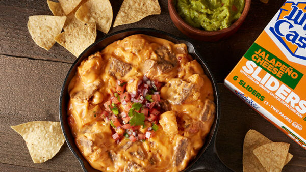 Bowl of queso dip next to a bowl of guacamole, a box of white castle jalapeno cheese sliders all surrounded by tortilla chips