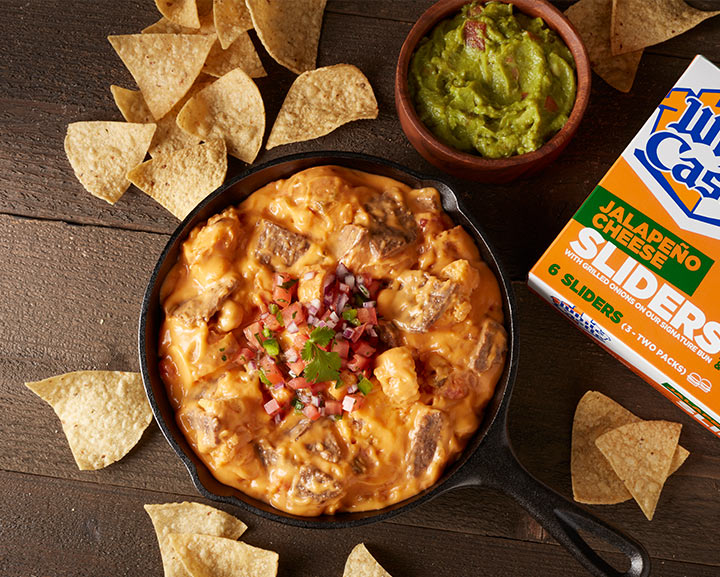 Bowl of queso dip next to a bowl of guacamole, a box of white castle jalapeno cheese sliders all surrounded by tortilla chips