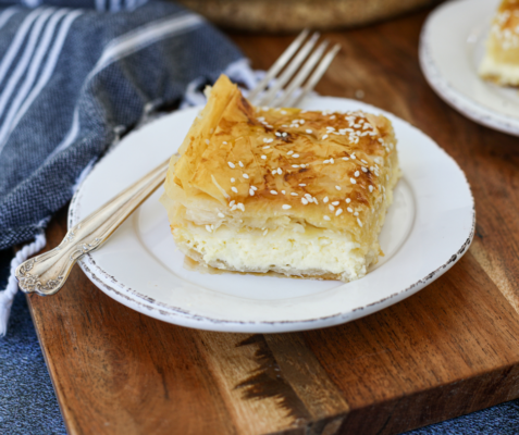 White plate with a fork and a slice of tiropita on top of it sitting on a wooden cutting board