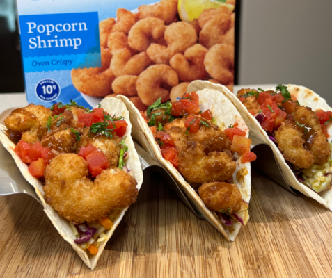 Close up of 3 fish tacos side by side and a box of seapak frozen popcorn shrimp in the background