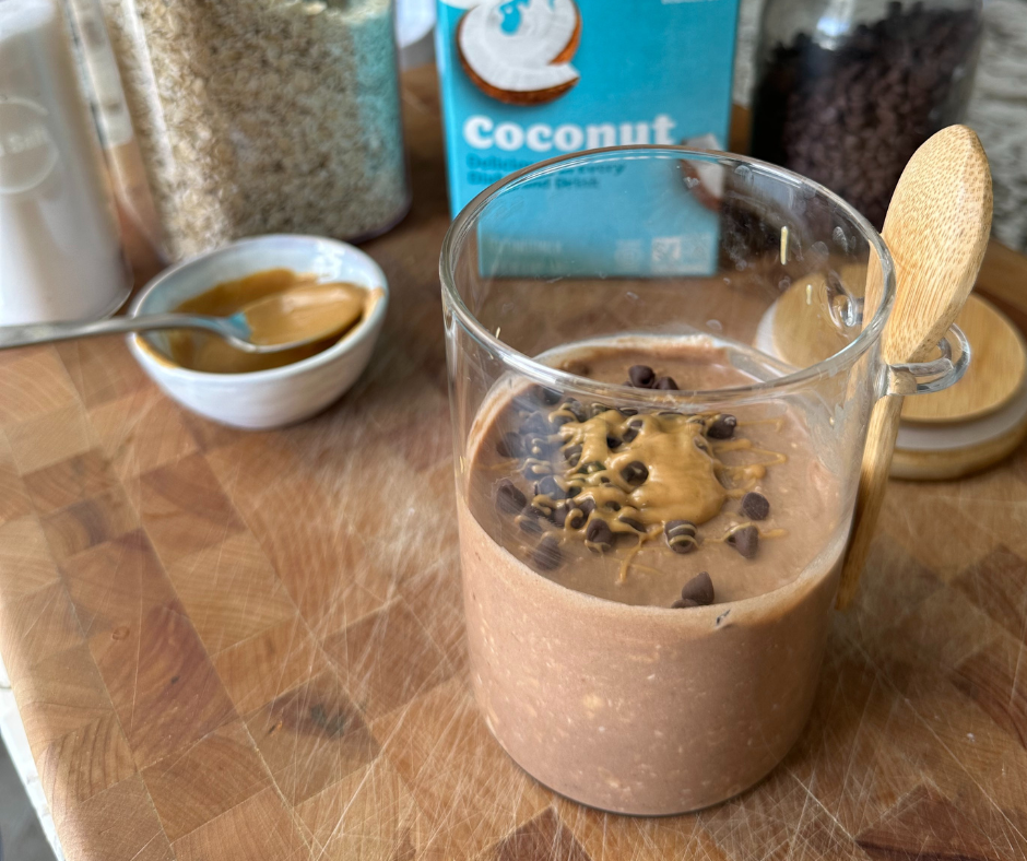A small glass with a spoon attached and filled with peanut butter overnight oats topped with chocolate chips and peanut butter and sitting on top of wooden cutting board with a blue carton of silk milk behind it