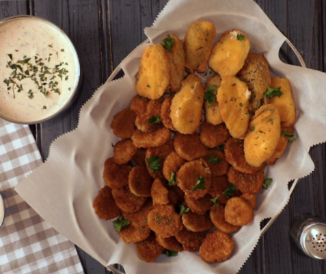 A bowl of fried pickles and jalapeno poppers on top of a wooden table, a bowl of southwest style ranch, and a gingham print placemat