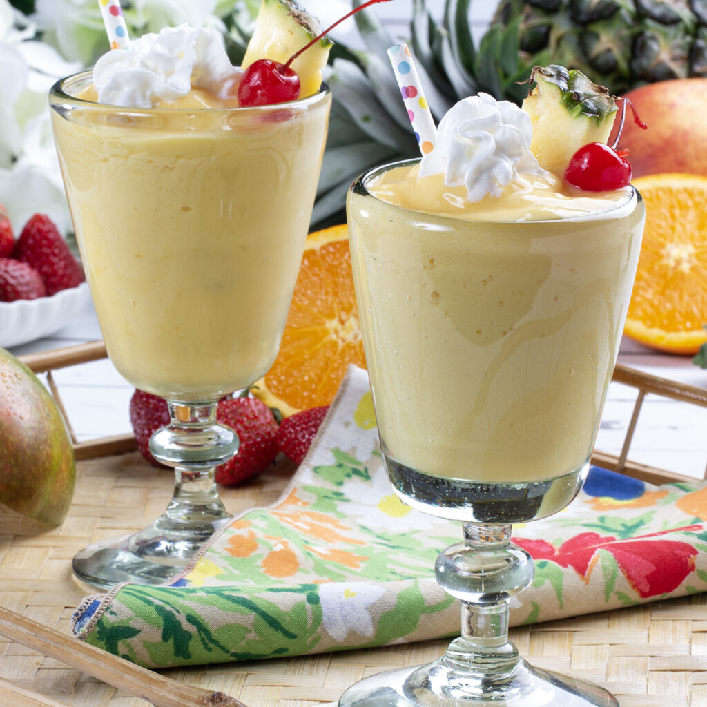 Two glasses filled with yellow blended beverage garnished with whip cream, pineapple, and a cherry