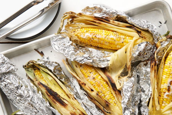 Four grilled stalks of corn wrapped in tin foil on a pan