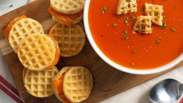 Mini waffle grilled cheeses on a cutting board next to a bowl of tomato soup