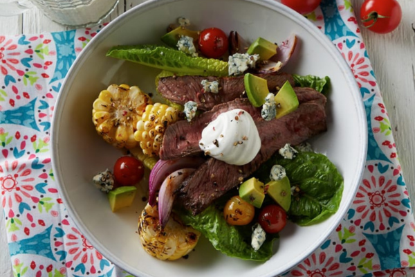 Grilled Steak, corn, tomatoes, avocados, and blue cheese in a white bowl sitting next to tomatoes on the vine on the right and a cup of ranch on the left