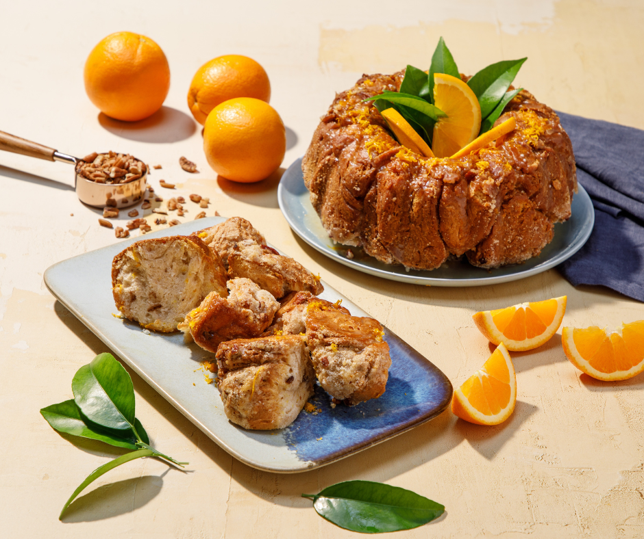 A close up shot of a plate of honey orange pull apart sweet bread with oranges, a loaf of honey orange pull apart sweet bread, and oranges besides it