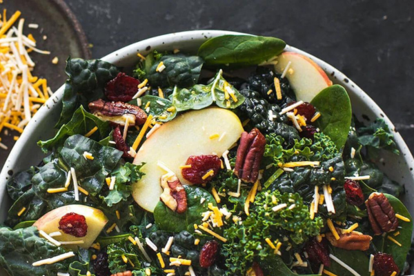 Close up shot of a white bowl filled with Kale Apple Cheddar Salad with an out of focus plate of shredded cheddar cheese in the background