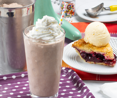 tall glass filled with a chocolate milkshake topped with whipped cream in front of a white plate with pie on top