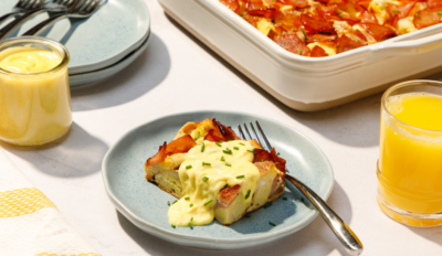 Zoomed out shot of a piece of overnight eggs benedict casserole on a plate with a fork besides it and 2 glasses of orange juice on each side, a pan of the casserole in the distance and a stack of plates with forks on top next to it