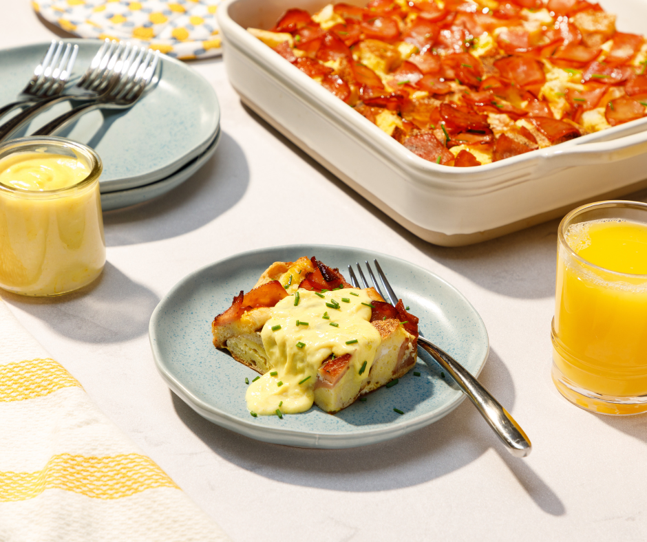 Zoomed out shot of a piece of overnight eggs benedict casserole on a plate with a fork besides it and 2 glasses of orange juice on each side, a pan of the casserole in the distance and a stack of plates with forks on top next to it