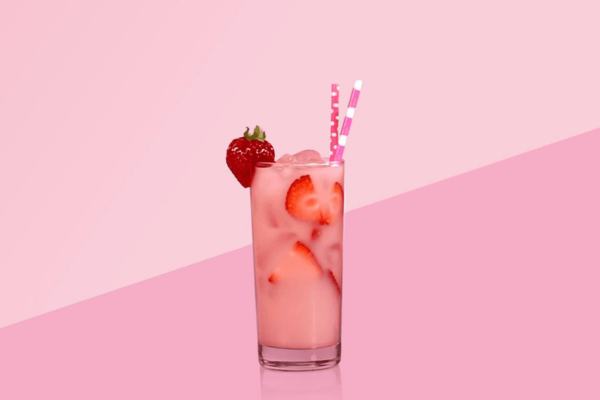 Tall glass of silk pink drink with two pink straws inside and a strawberry on the rim against a pink background