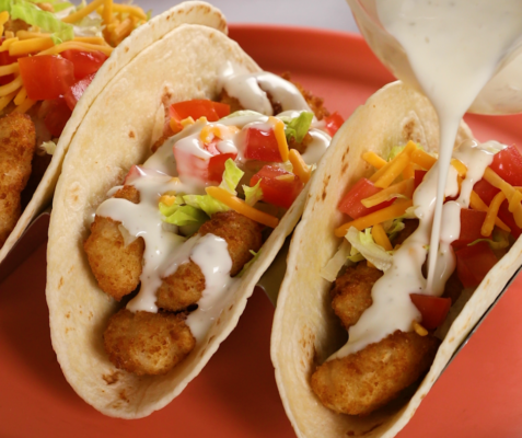 Two tacos side by side, one being drizzled with a creamy sauce