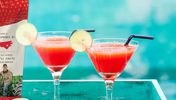 Two margarita glasses filled with a strawberry margarita and a cucumber on the rim glasses sitting on a table 