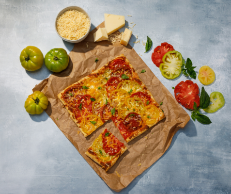 Overhead shot of three cheese summer tomato tart surrounded by green and red tomatoes and a bowl of cheese