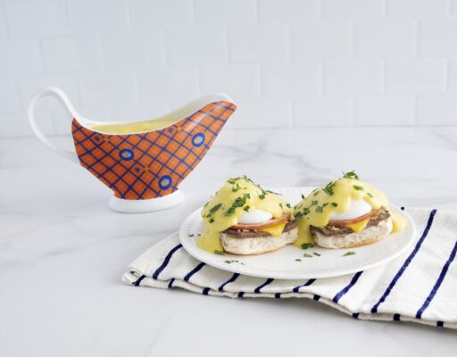 Eggs benedict on a white plate sitting on top of a black and white striped cloth and a gravy boat of hollandaise sauce next to it