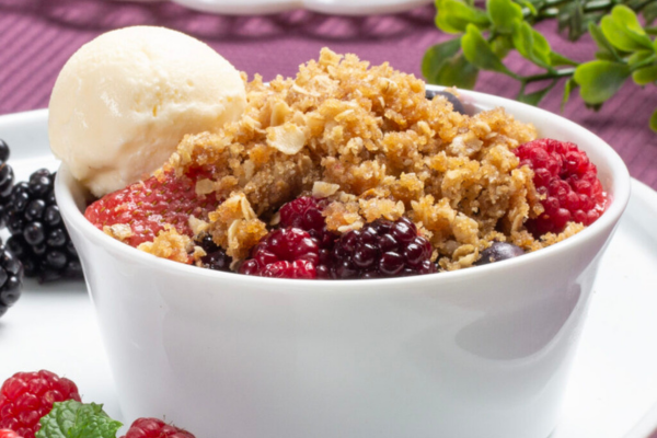close up shot of a white bowl filled with berries topped with crumble and vanilla iced cream