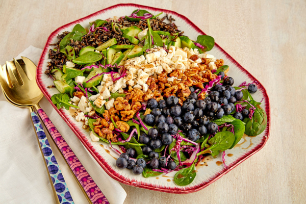 Close up image of a white rectangular dish with red edges sitting next to a fork and spoon and filled with seperated lines of spinach, blueberries, walnuts, feta, avocado, and wild rice.