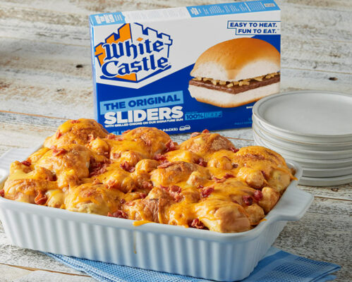 baking dish filled with mini cheeseburgers with cheese and tater tots sprinkled on top with a box of white castle original sliders in the background and white dishes to the right